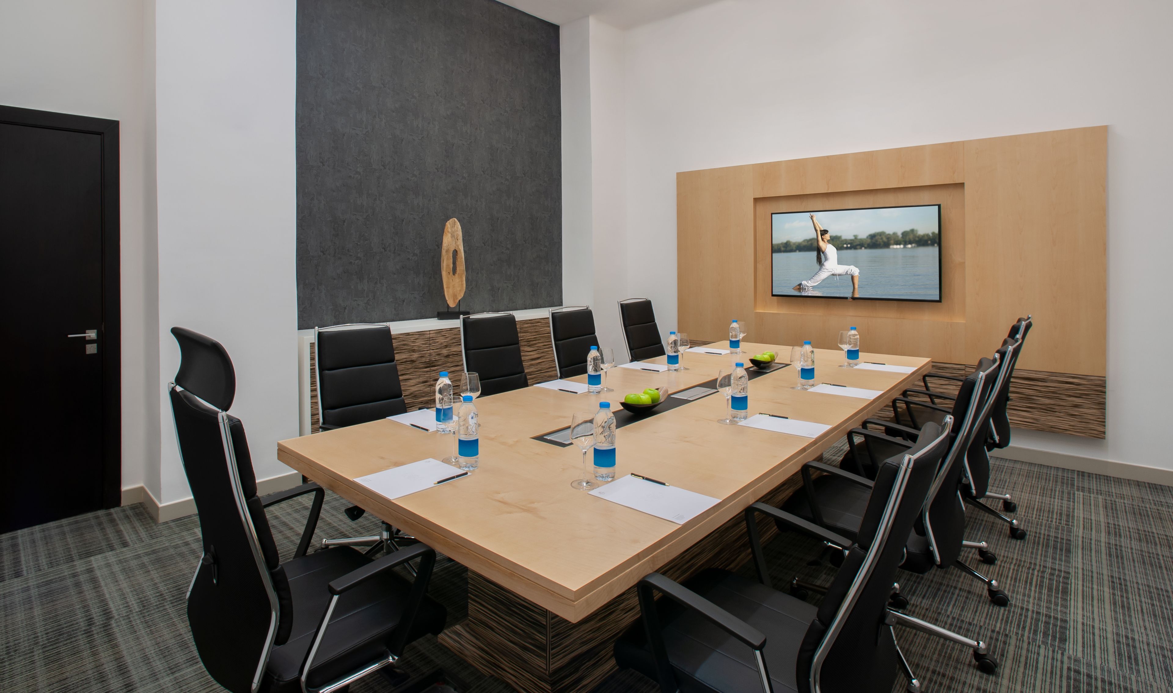 Ein Konferenzraum mit einem langen Tisch, schwarzen Stühlen, Wasserflaschen und einem Fernseher an der Wand, der eine Yoga-Pose zeigt.