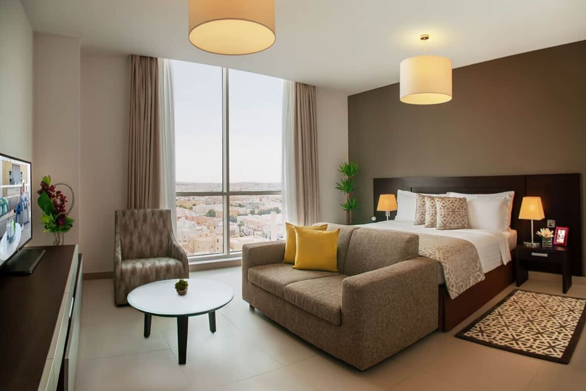 A hotel room with a large window, a sofa, a bed with patterned pillows, a small table, and a TV on a stand.