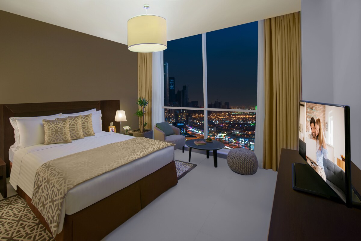 A hotel room with a large bed, a TV, a desk, and a view of a cityscape at night.