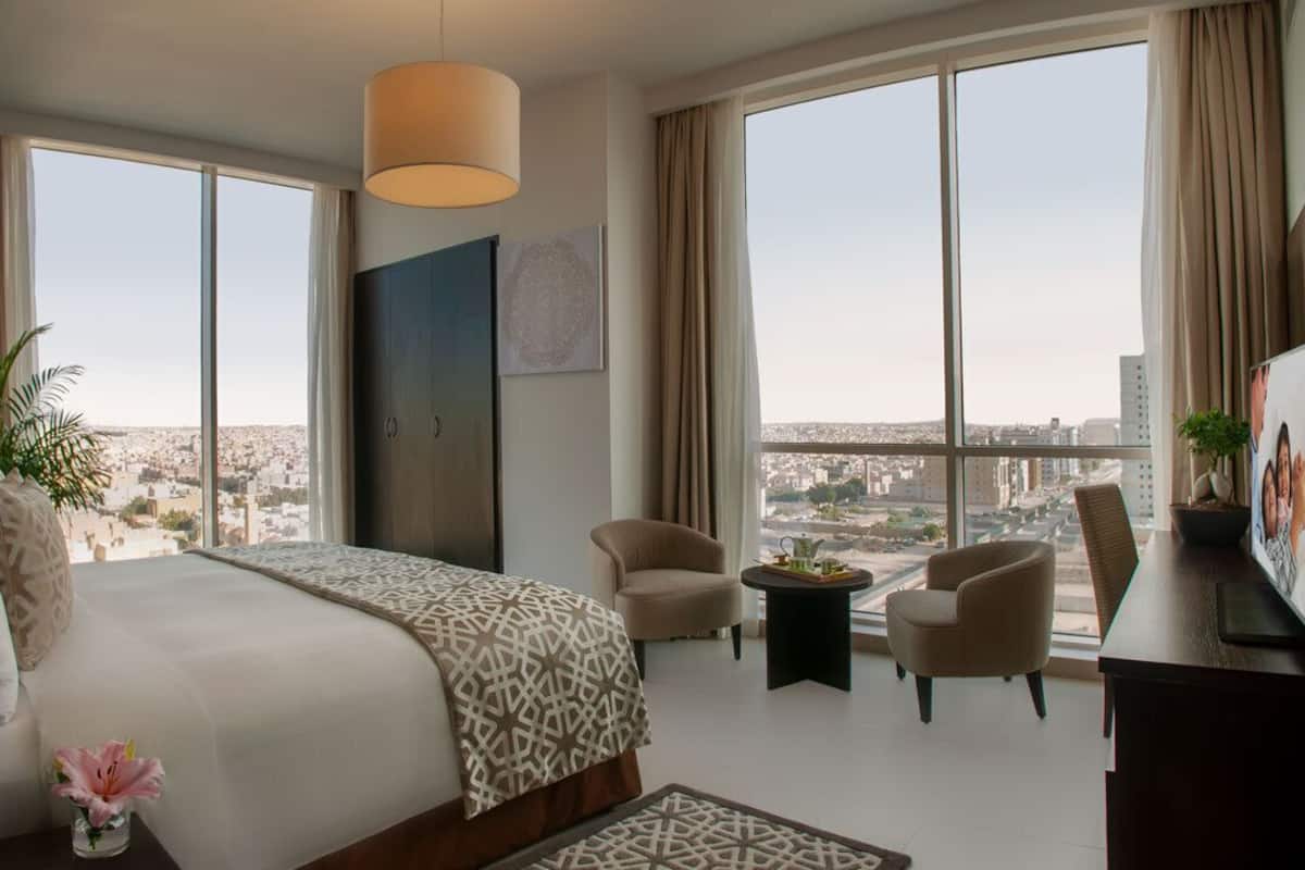 A spacious hotel room with a large bed, two armchairs, a small table, and a TV. The room has large windows offering a city view.