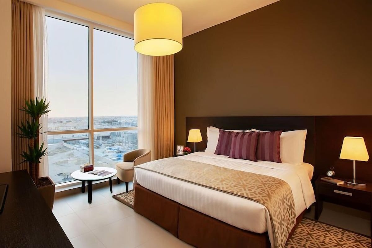 A hotel room with a large bed, two bedside lamps, a small round table, a chair, a TV, and a large window with a city view.