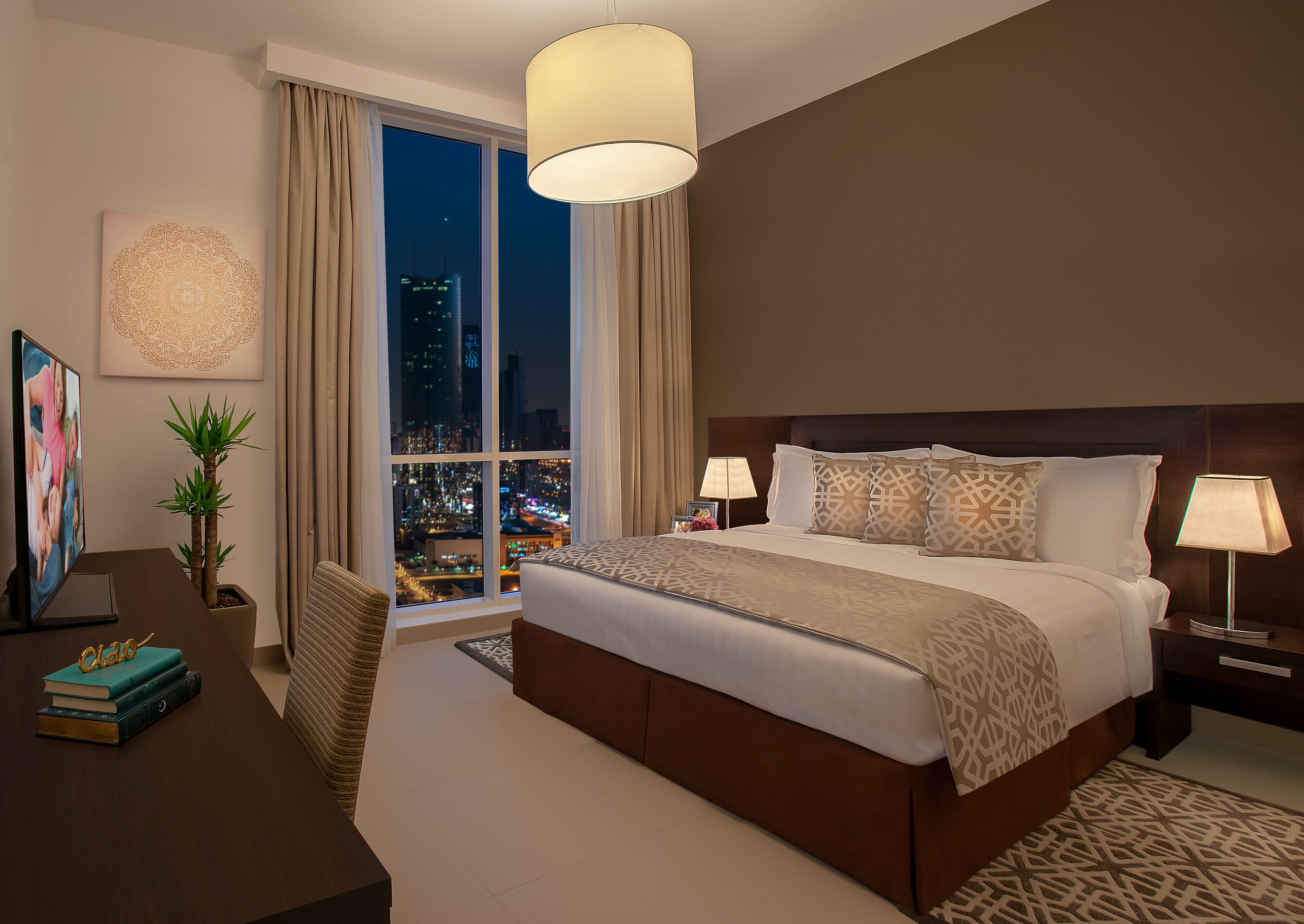 A modern hotel room with a large bed, two bedside lamps, a desk with a laptop, a potted plant, and a city view through large windows.