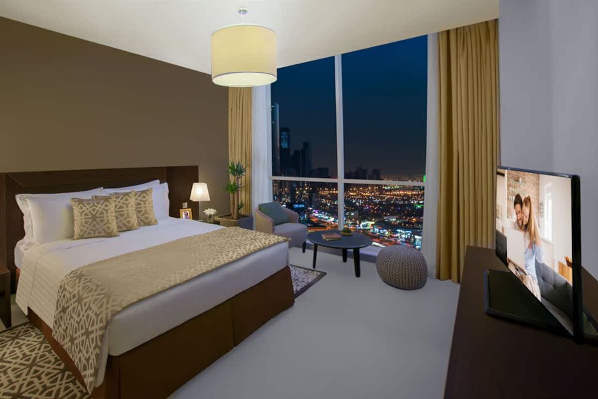A hotel room with a large bed, a TV, a desk, and a view of a cityscape at night.