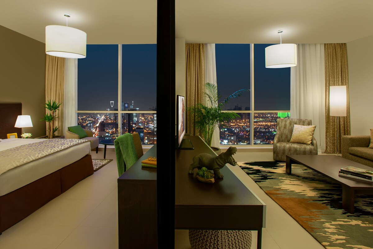 A hotel room with a large bed, a sofa, a coffee table, and a view of a cityscape at night.