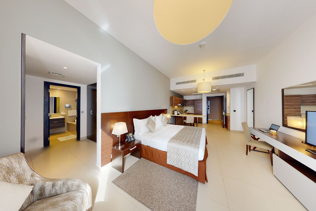 A modern hotel room with a large bed, a desk, a TV, and a separate living area.