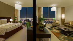 Ein Hotelzimmer mit einem großen Bett, einer Sitzecke inklusive Sofa und Sesseln, einem Couchtisch und Blick auf eine nächtliche Stadtlandschaft.