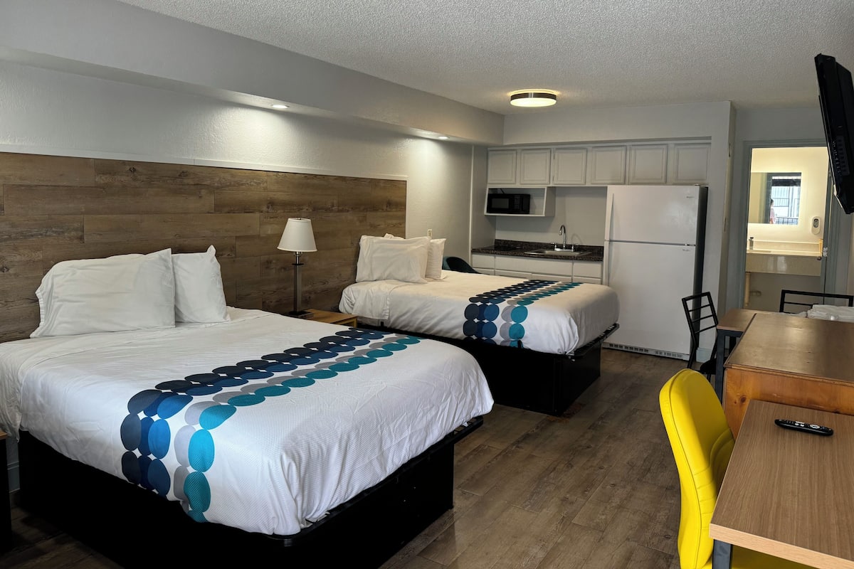 A hotel room with two beds, a desk, a chair, a microwave, a refrigerator, and a sink.