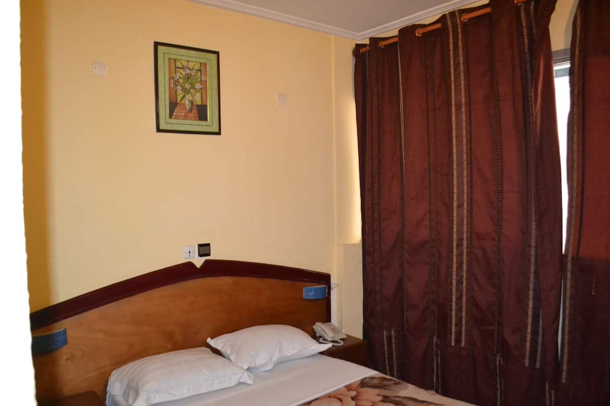A hotel room with a single bed, a nightstand with a phone, a wall-mounted light switch, and a framed picture above the bed.