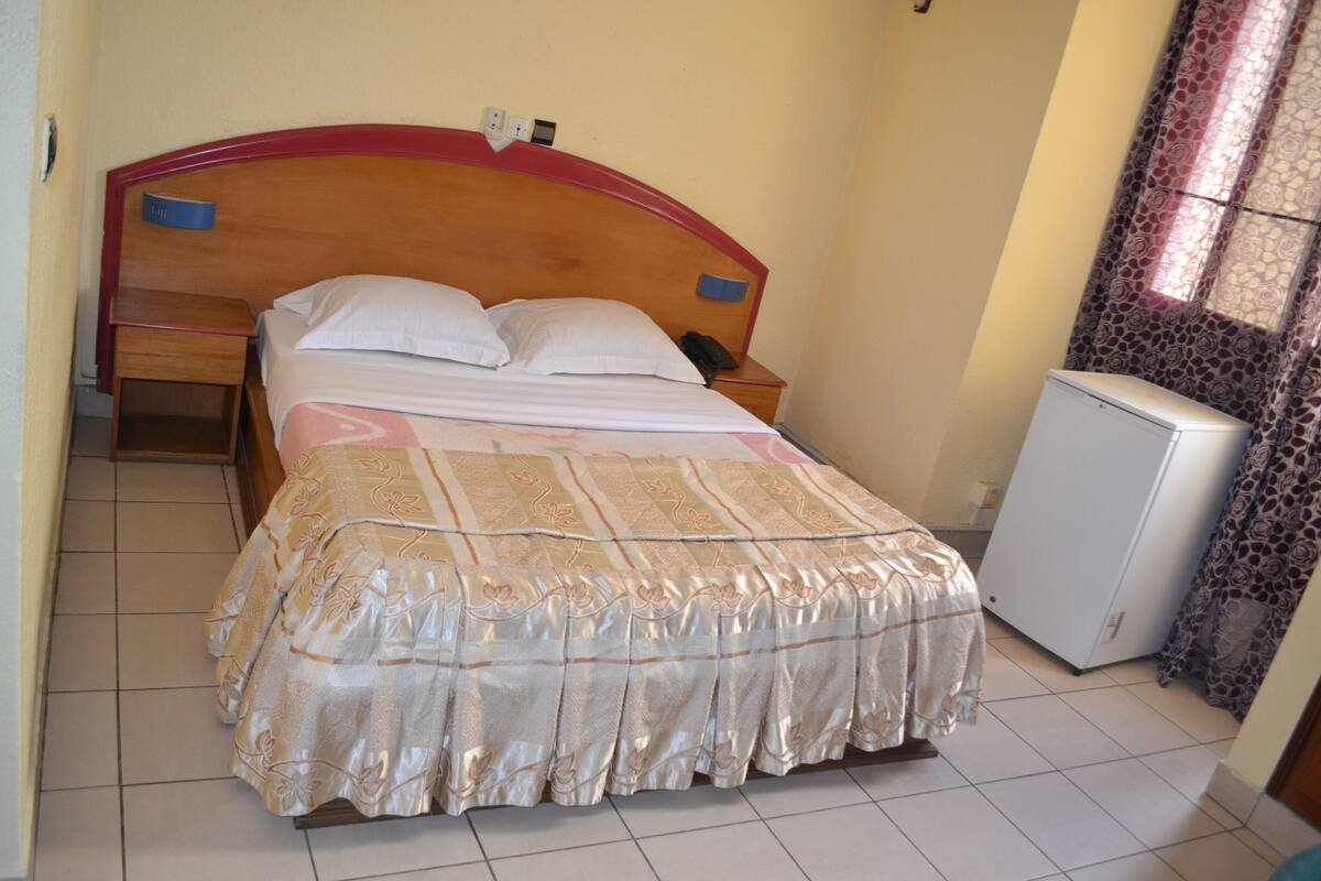 A bedroom with a wooden headboard, a bed with a patterned bedspread, a bedside table with a lamp, and a refrigerator.