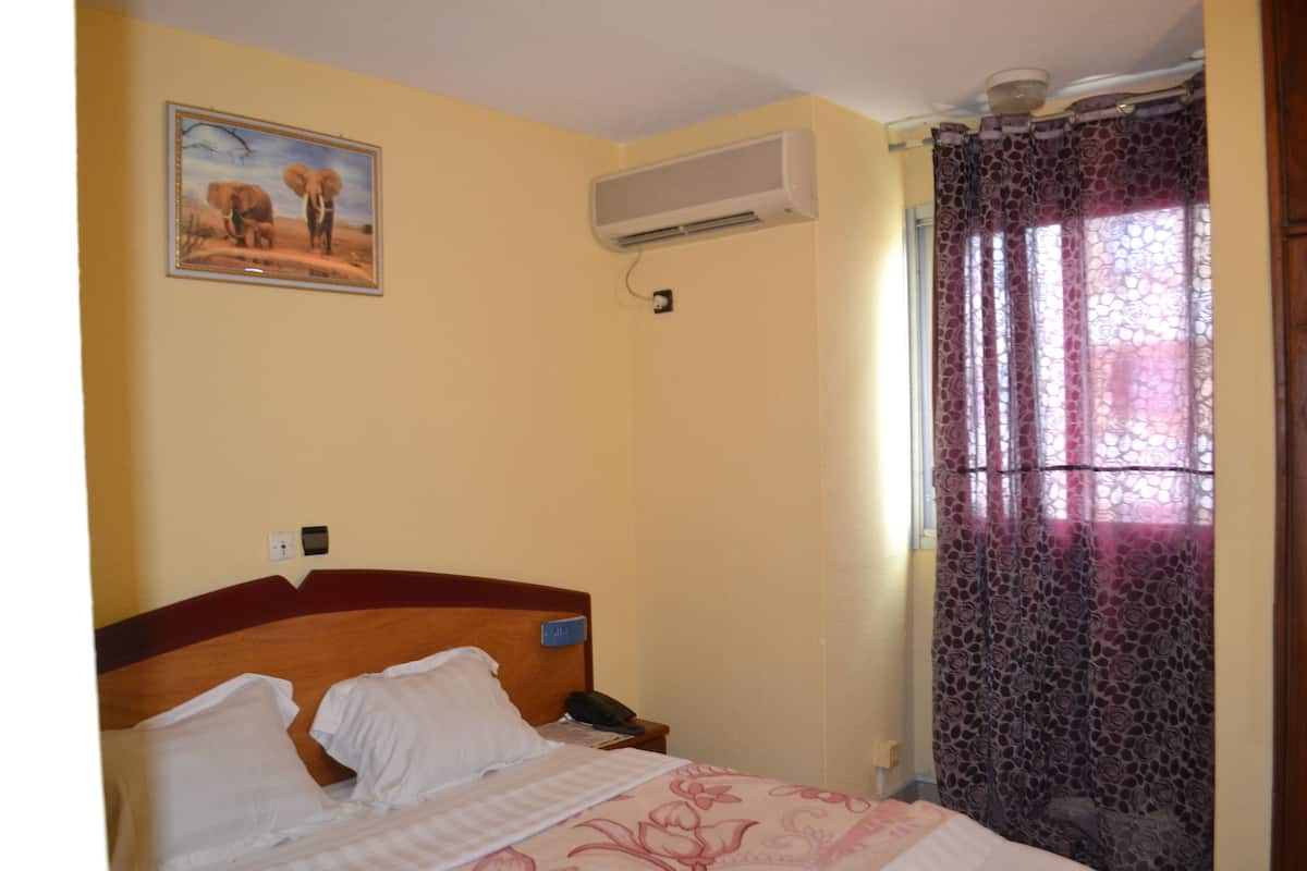 A hotel room with a bed, a wall-mounted air conditioner, a painting of elephants, and a window with a patterned curtain.