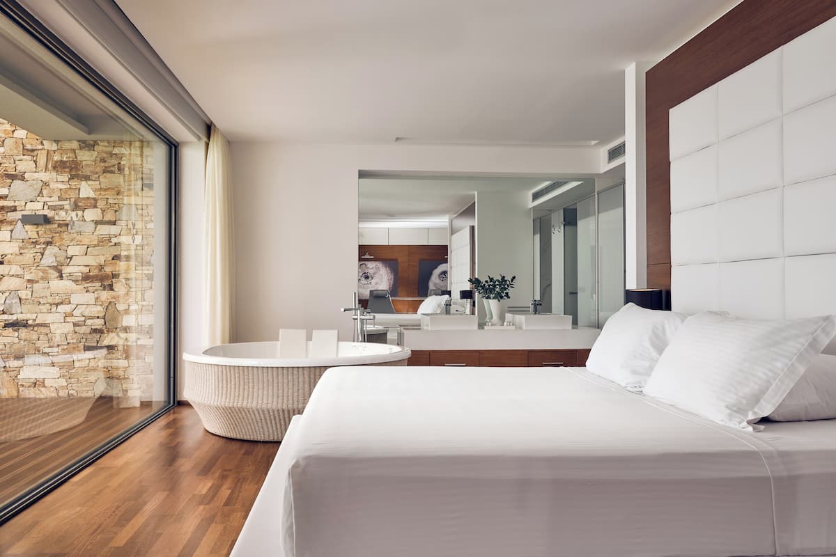 A modern hotel room with a large bed, a round seating area, a stone wall, and a large mirror.