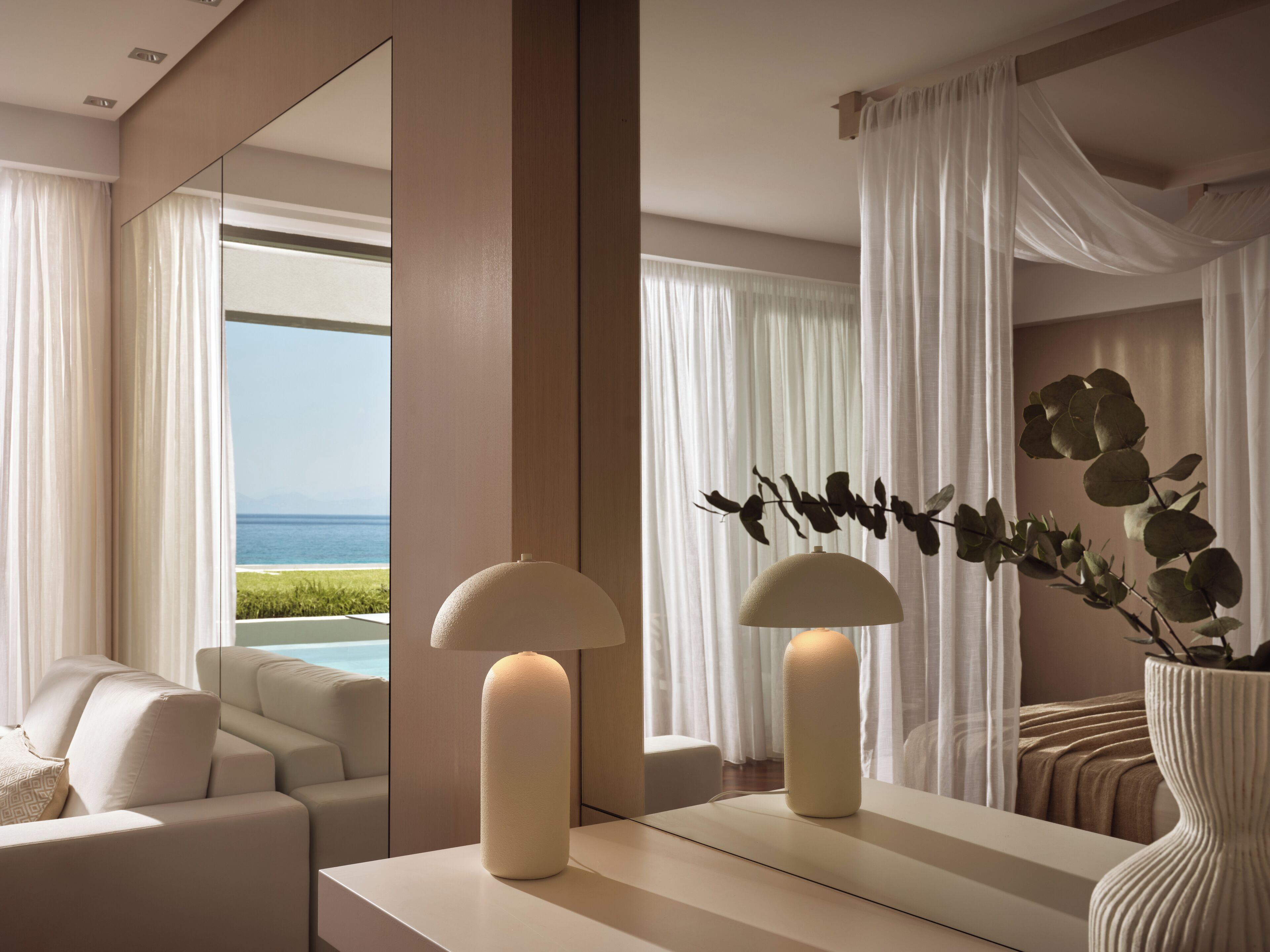 Habitación de hotel moderna con un espejo grande que refleja una vista a la playa, un sofá y un jarrón con plantas.