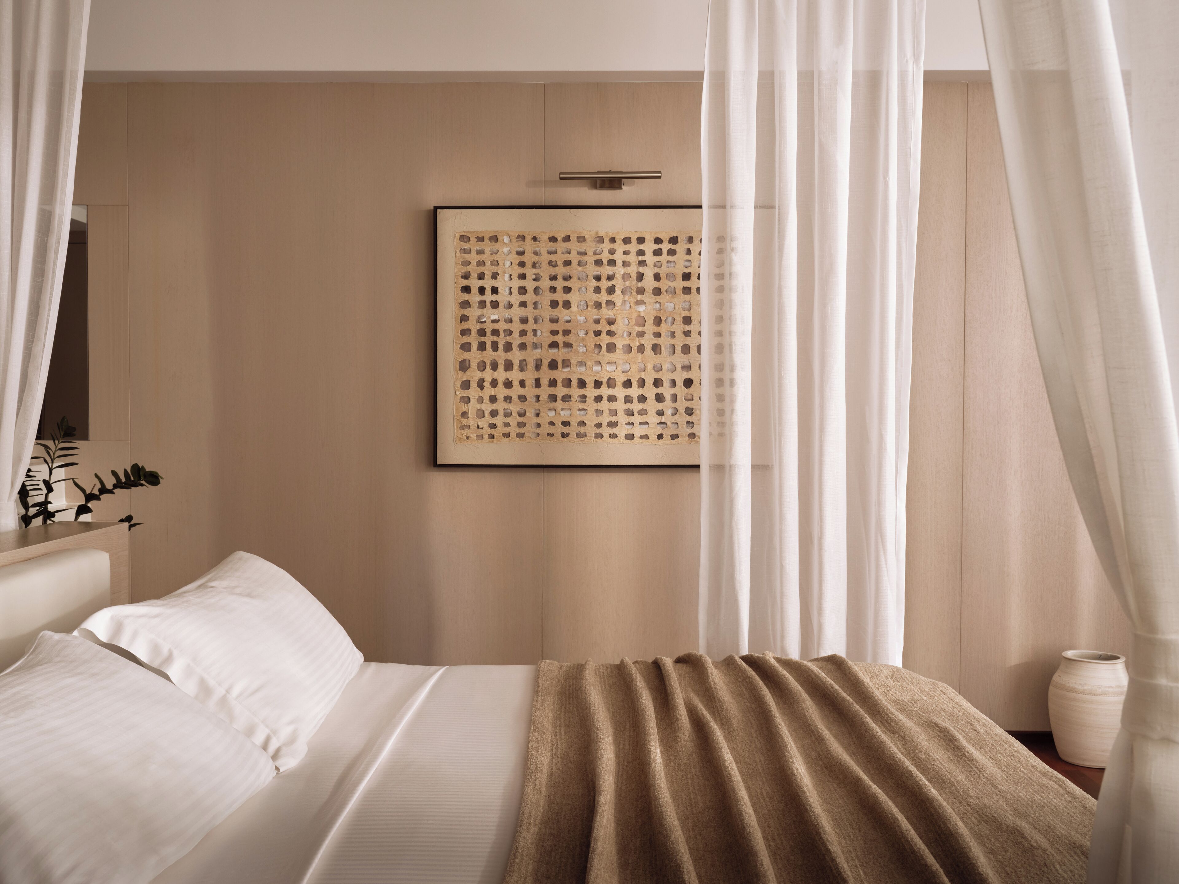 A bedroom with a bed, white curtains, a wall-mounted artwork, and a bedside table with a vase.