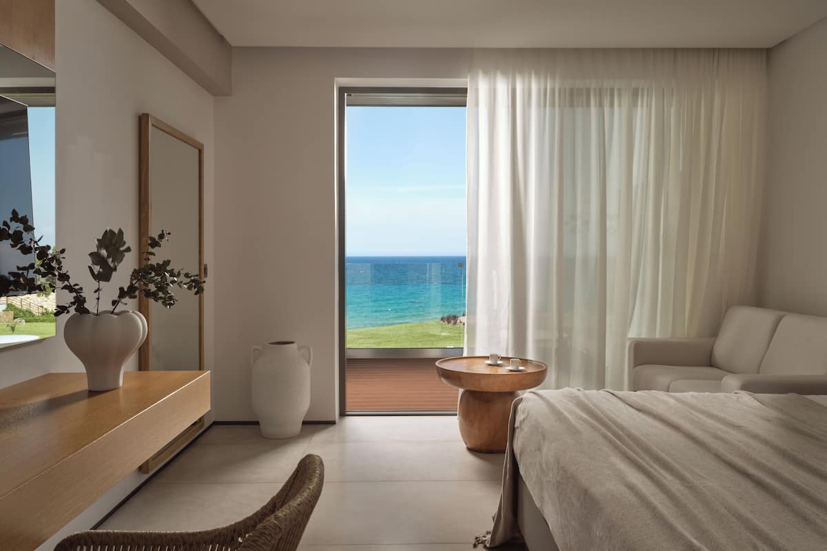 A modern bedroom with a large bed, a seating area, a small table, a potted plant, and a view of the ocean.