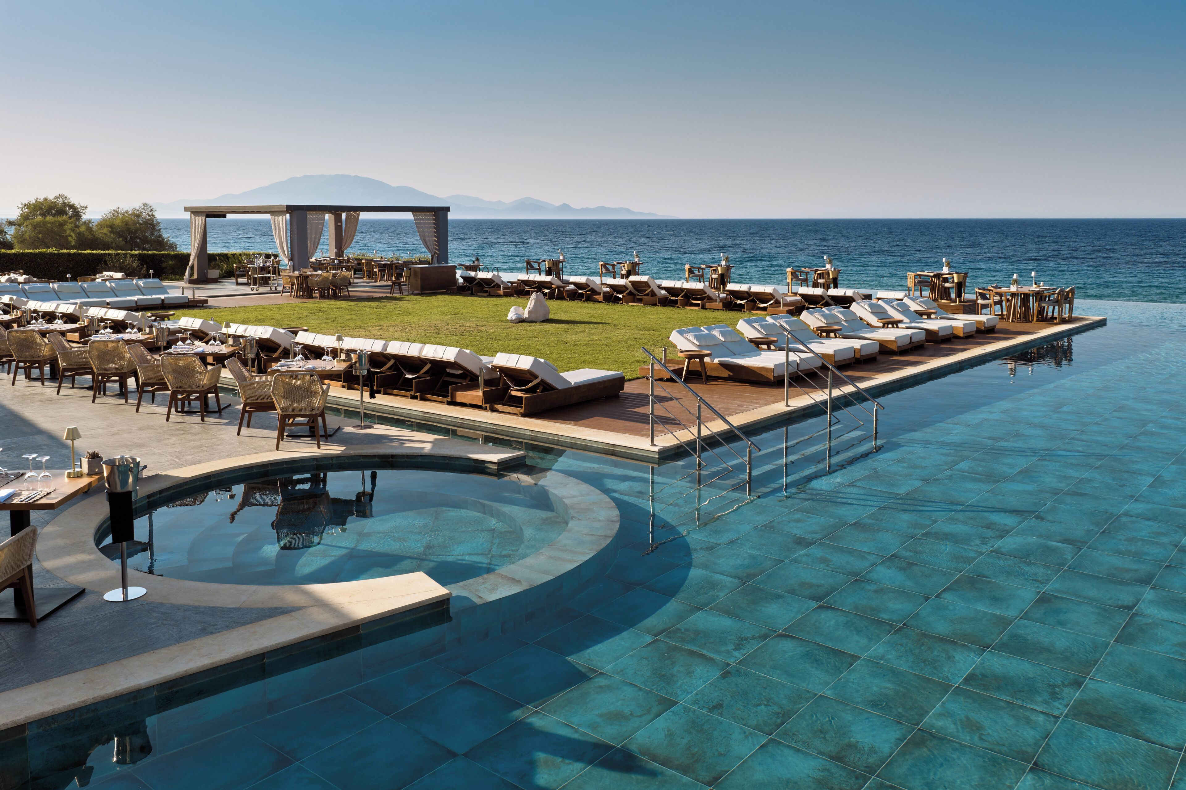A poolside area with lounge chairs, tables, and a dining setup overlooking the sea.