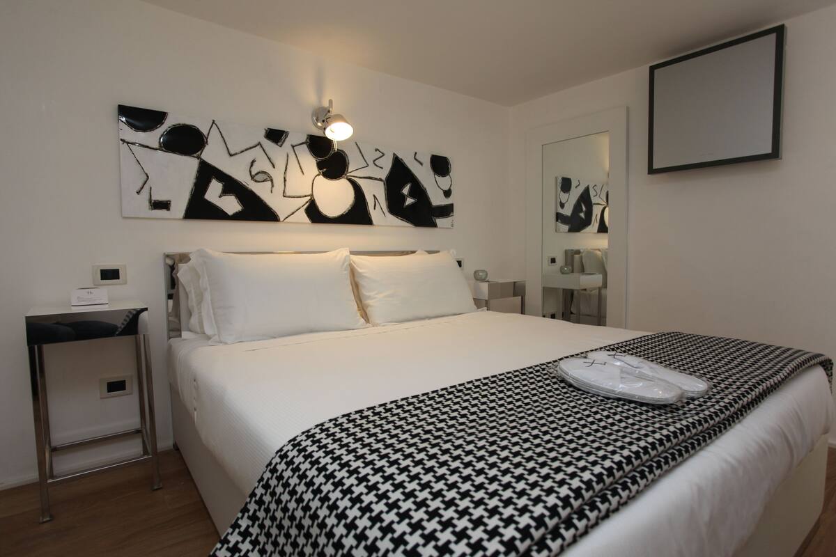Quarto de hotel com uma cama grande, mesa de cabeceira, luminária e uma obra de arte na parede, além de um espelho.
