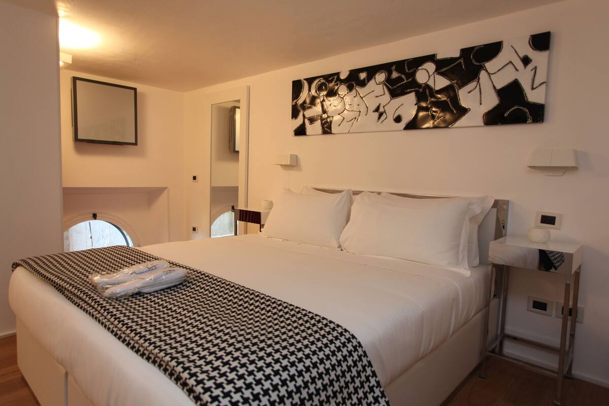 Quarto de hotel com uma cama grande, manta xadrez houndstooth, pintura abstrata moderna, mesa de cabeceira com abajur e espelho na parede.