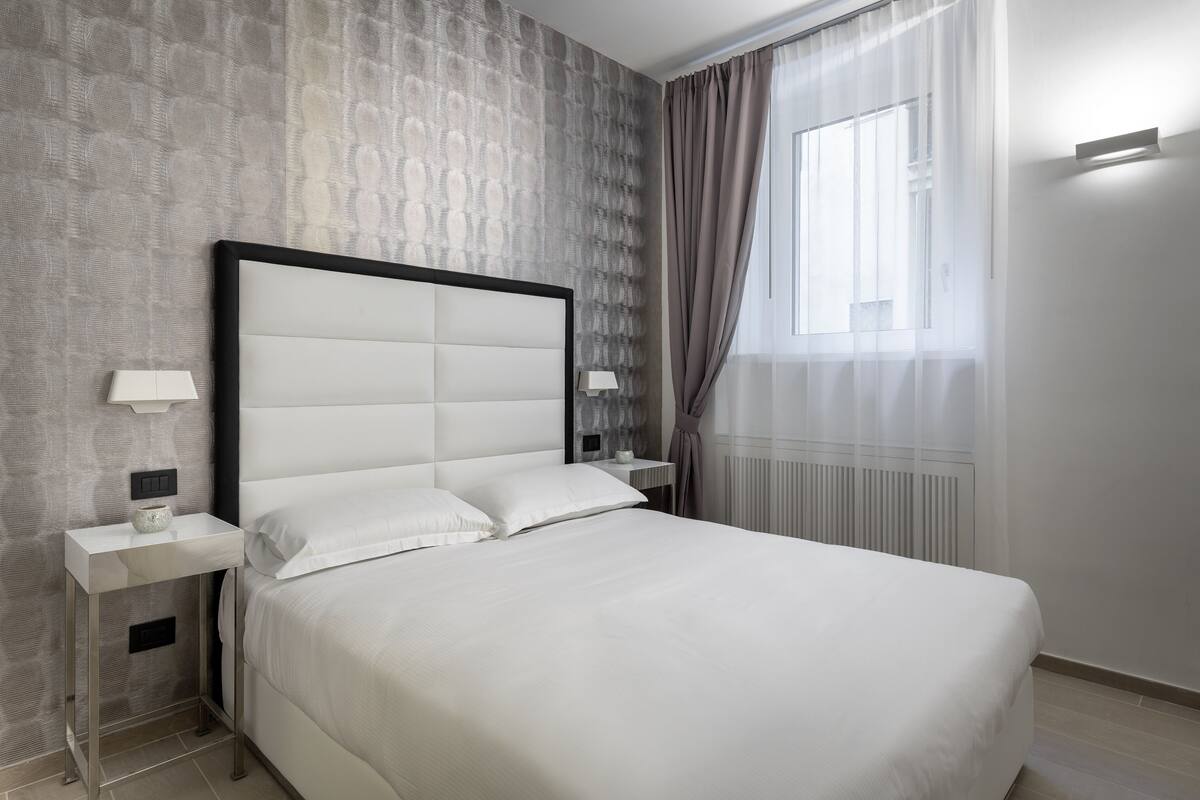 Quarto de hotel moderno com uma cama grande, uma mesinha de cabeceira com abajur e uma janela com cortinas.