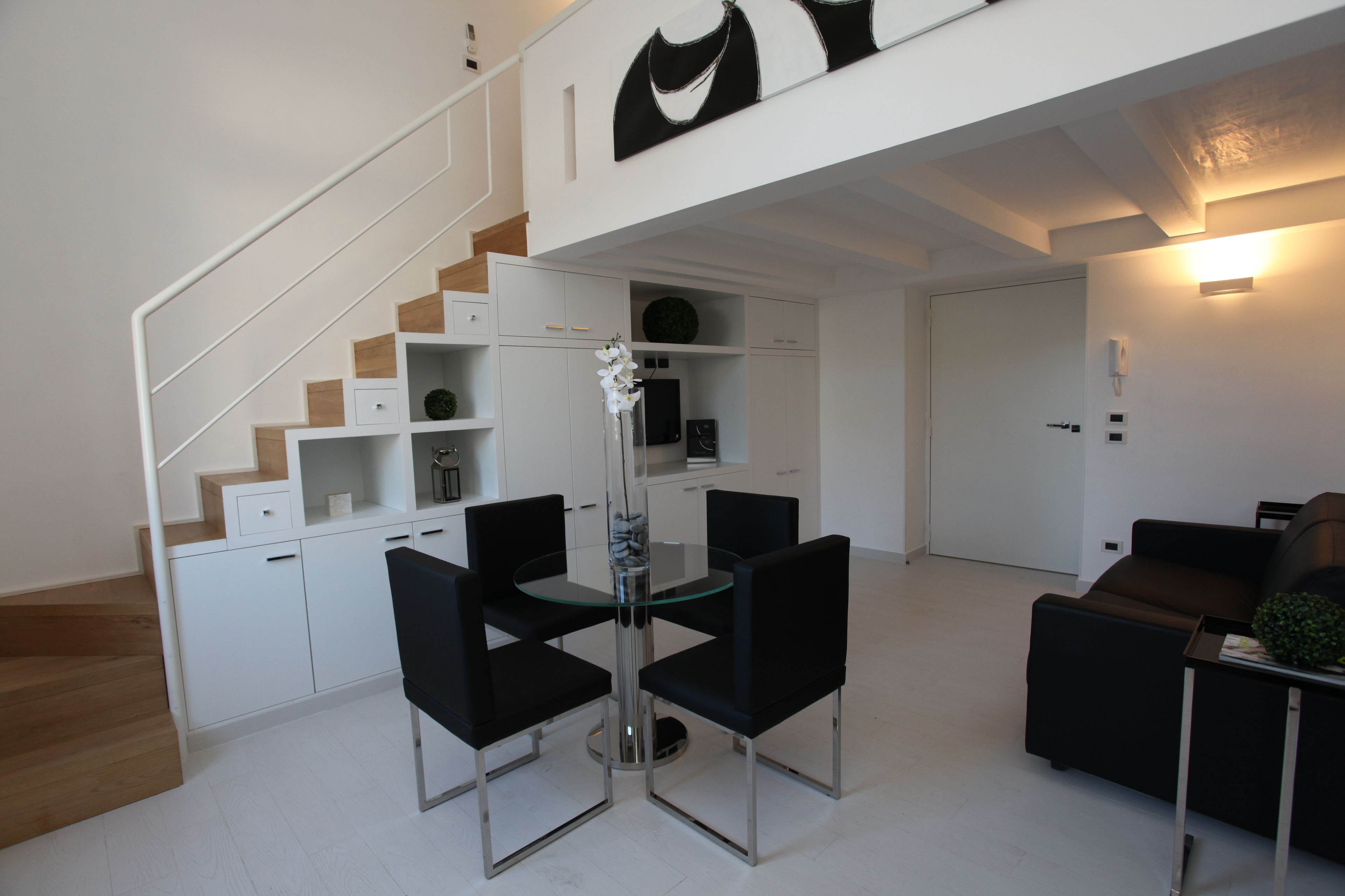 A modern living room with a glass-top dining table, black chairs, a brown sofa, and a staircase with white cabinets.