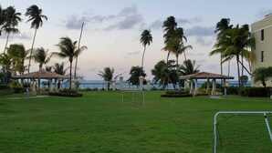 Zona verde con un arco de fútbol, palmeras y dos pabellones junto a una masa de agua.