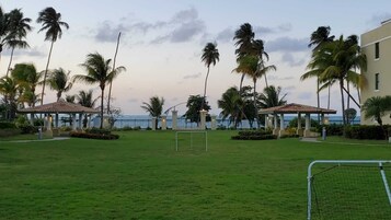 Zona verde con un arco de fútbol, palmeras y dos pabellones junto a una masa de agua.