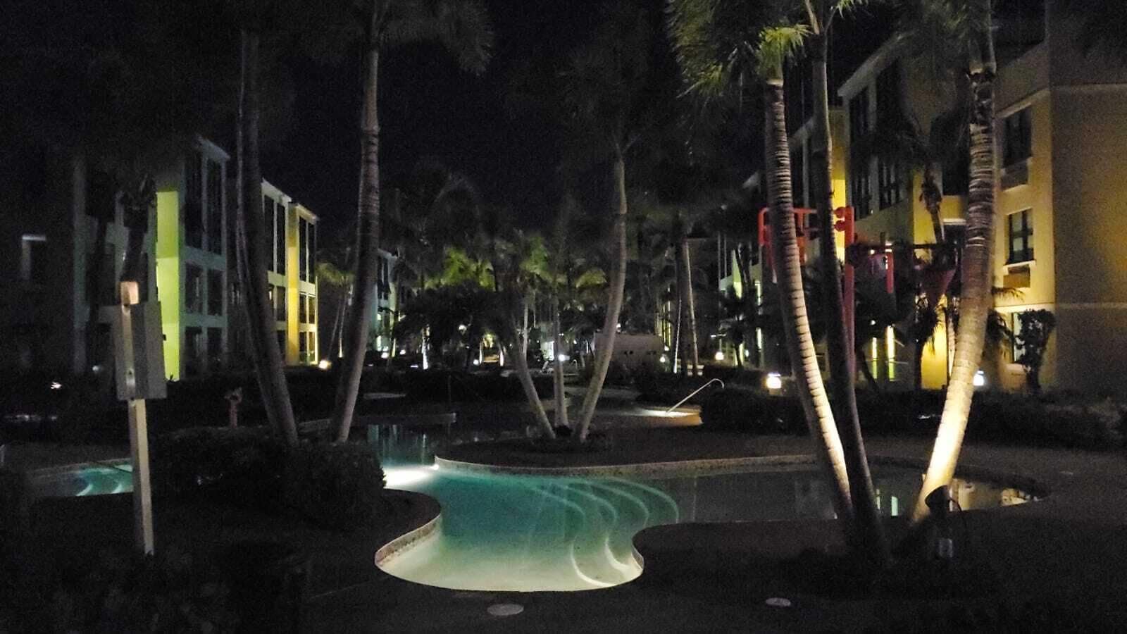 A nighttime view of a resort area with a swimming pool, palm trees, and illuminated buildings.