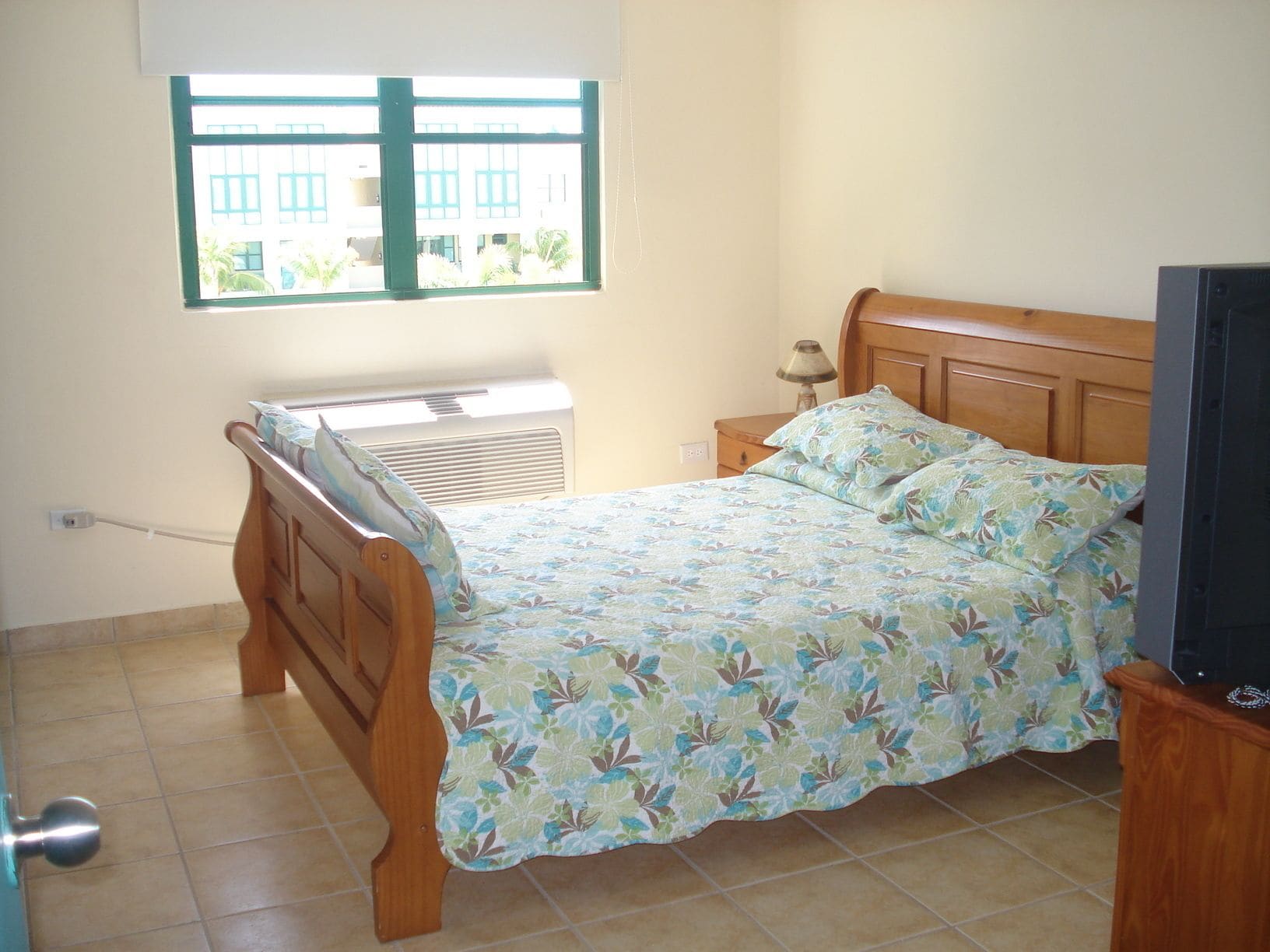 A bedroom with a bed, a window, an air conditioner, a nightstand, and a television.