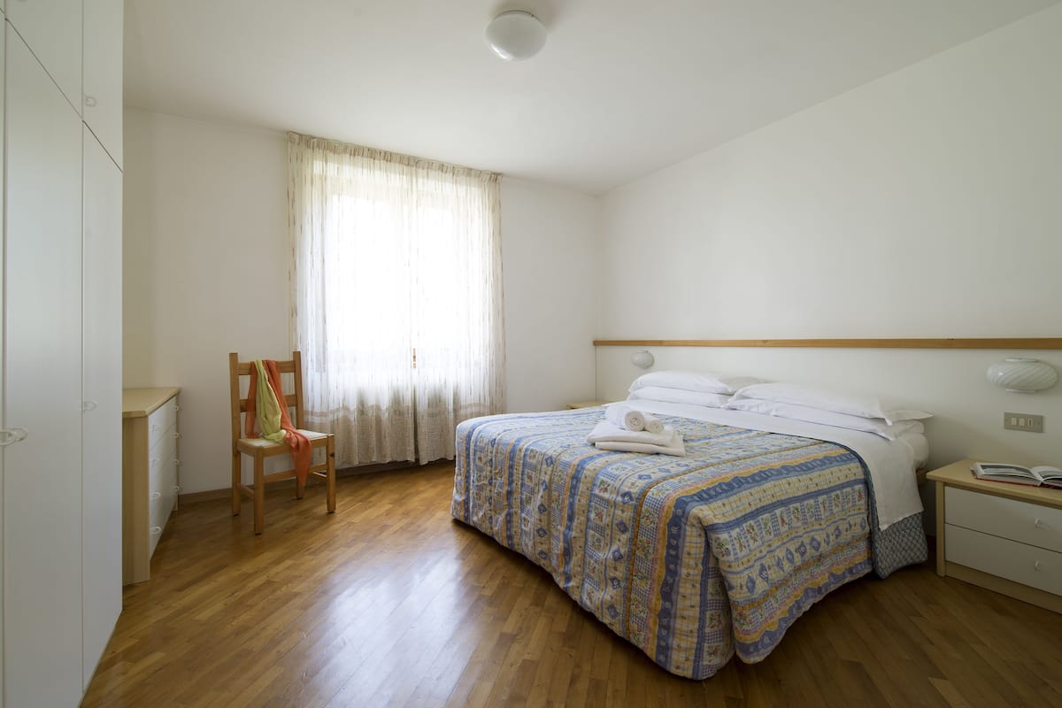 Ein Schlafzimmer mit Holzboden, einem Bett mit gemustertem Überwurf, einem Sessel, einem Nachttisch mit Lampe und einem Fenster mit Vorhängen.