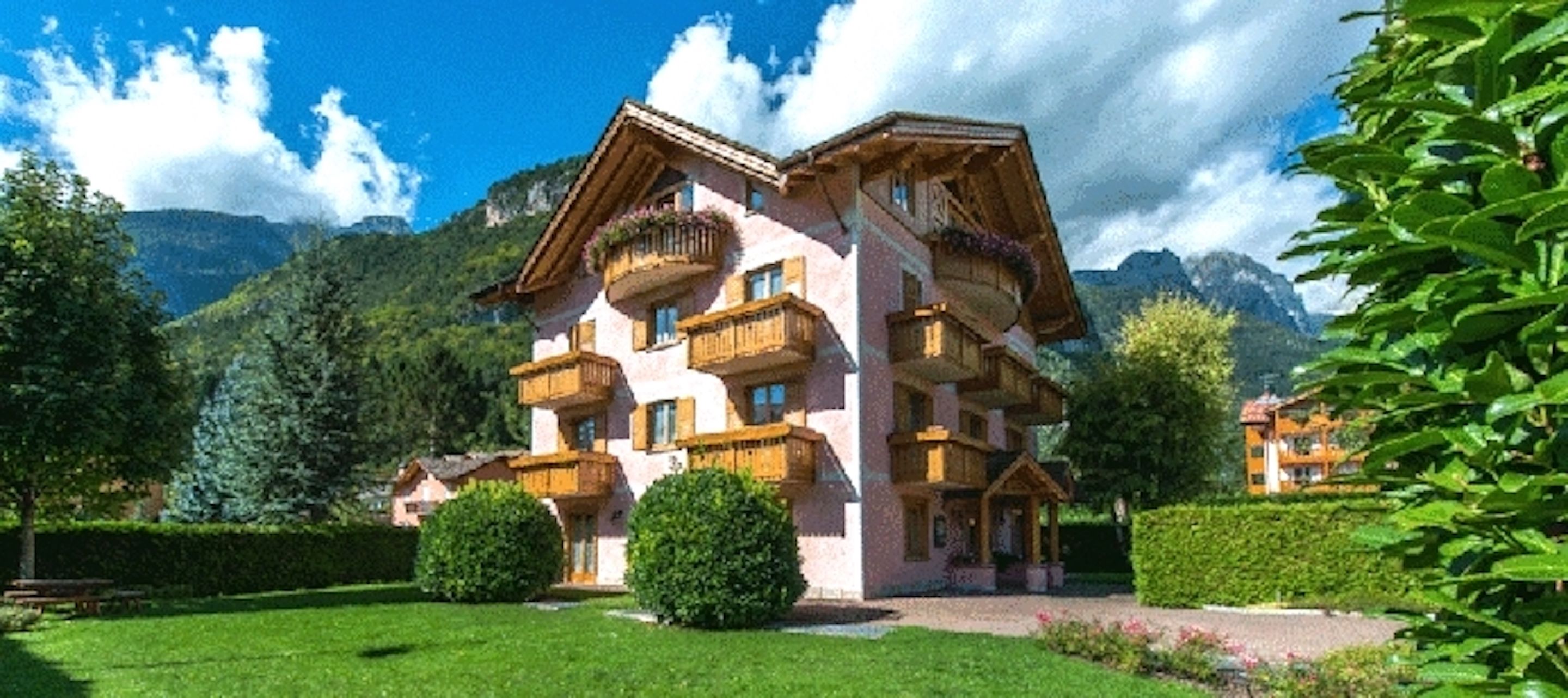 A multi-story chalet with balconies, surrounded by lush greenery and mountains.