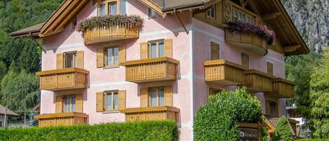 A multi-story building with wooden balconies and flower boxes, surrounded by greenery and a mountain backdrop.