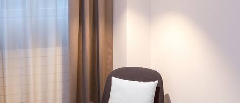 A modern hotel room with a dark brown armchair, a white pillow, a small table with a cup and book, and sheer curtains.