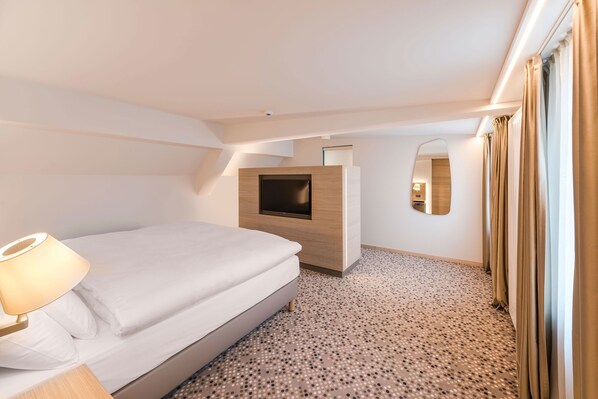 Ein modernes Hotelzimmer mit Bett, Fernseher an einer Holzvertäfelung, einem kleinen Nachttisch mit Lampe und einem gemusterten Teppich.