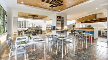 A spacious dining area with multiple tables and chairs, a kitchen area with modern appliances, and a view of high-rise buildings.