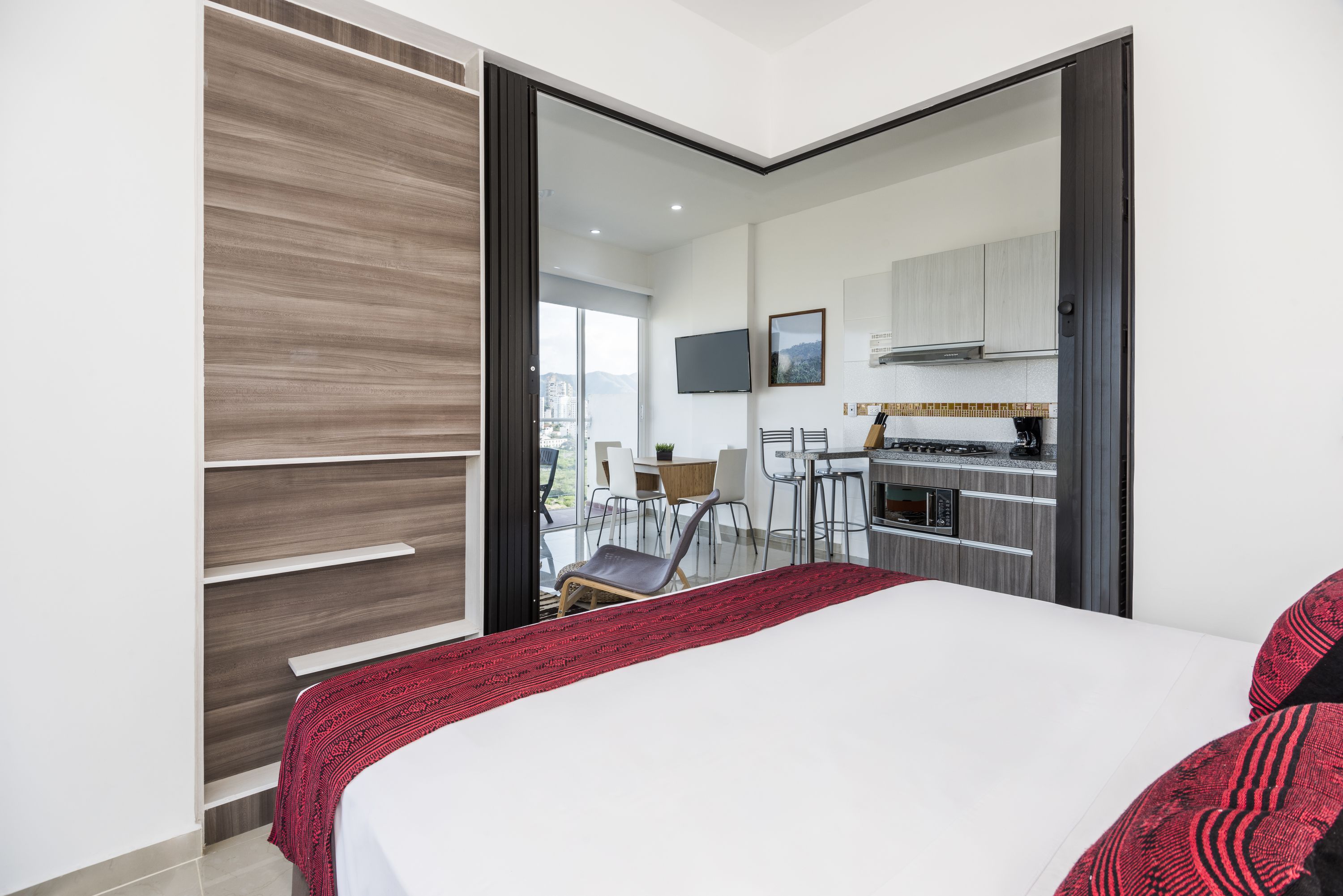 Een moderne hotelkamer met een groot bed, een rode gebogen deken, een keuken met een fornuis en stoelen, en een schuifdeur naar een balkon met uitzicht op de stad.