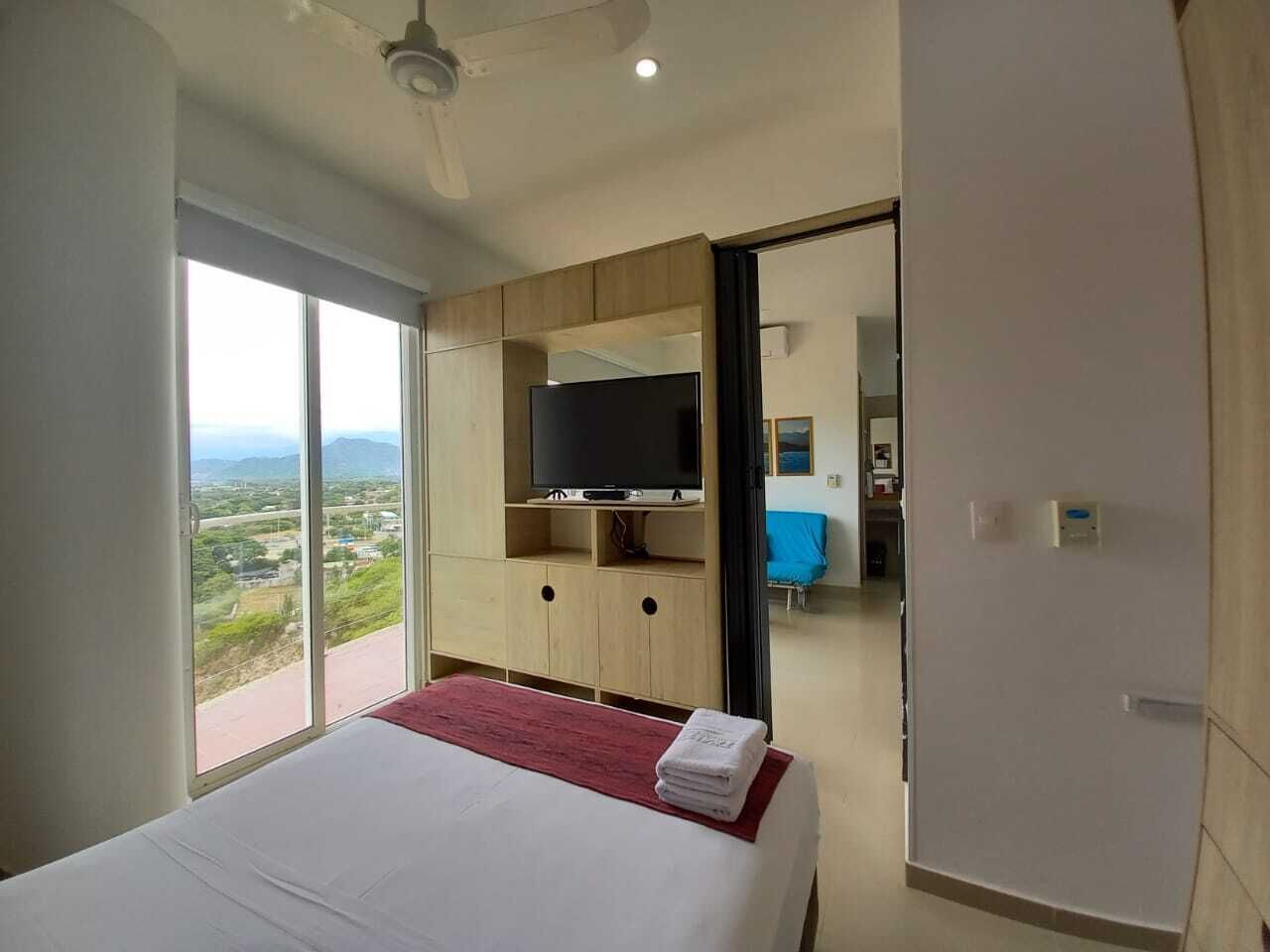 A hotel room with a large bed, a TV on a wooden cabinet, a balcony with a view, and a sliding door to a bathroom.
