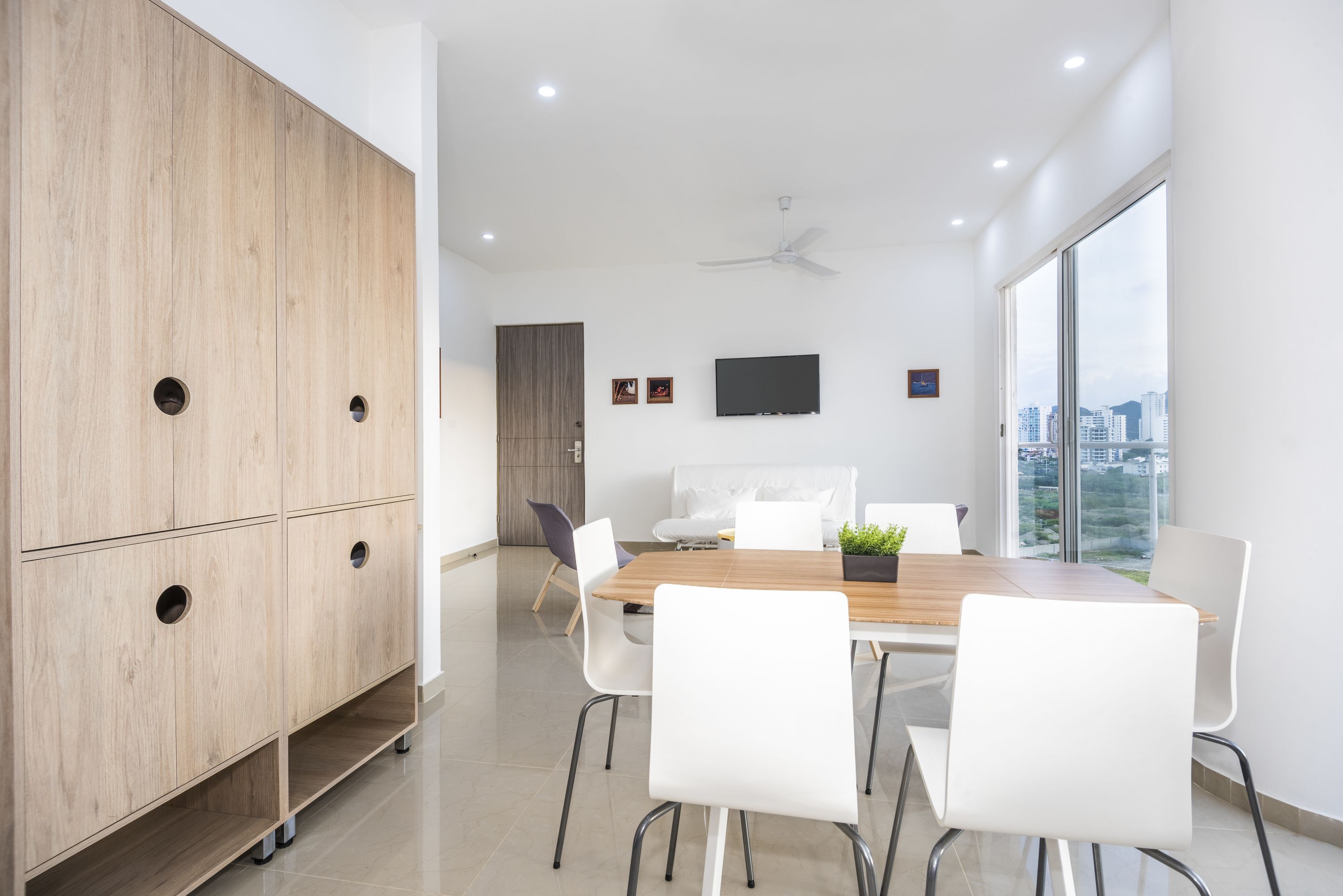 Una sala moderna con un aparador de madera, una mesa de comedor con sillas blancas, un sofá y un televisor montado en la pared.
