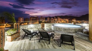 A rooftop terrace with a hot tub, lounge chairs, and a city skyline view at dusk.
