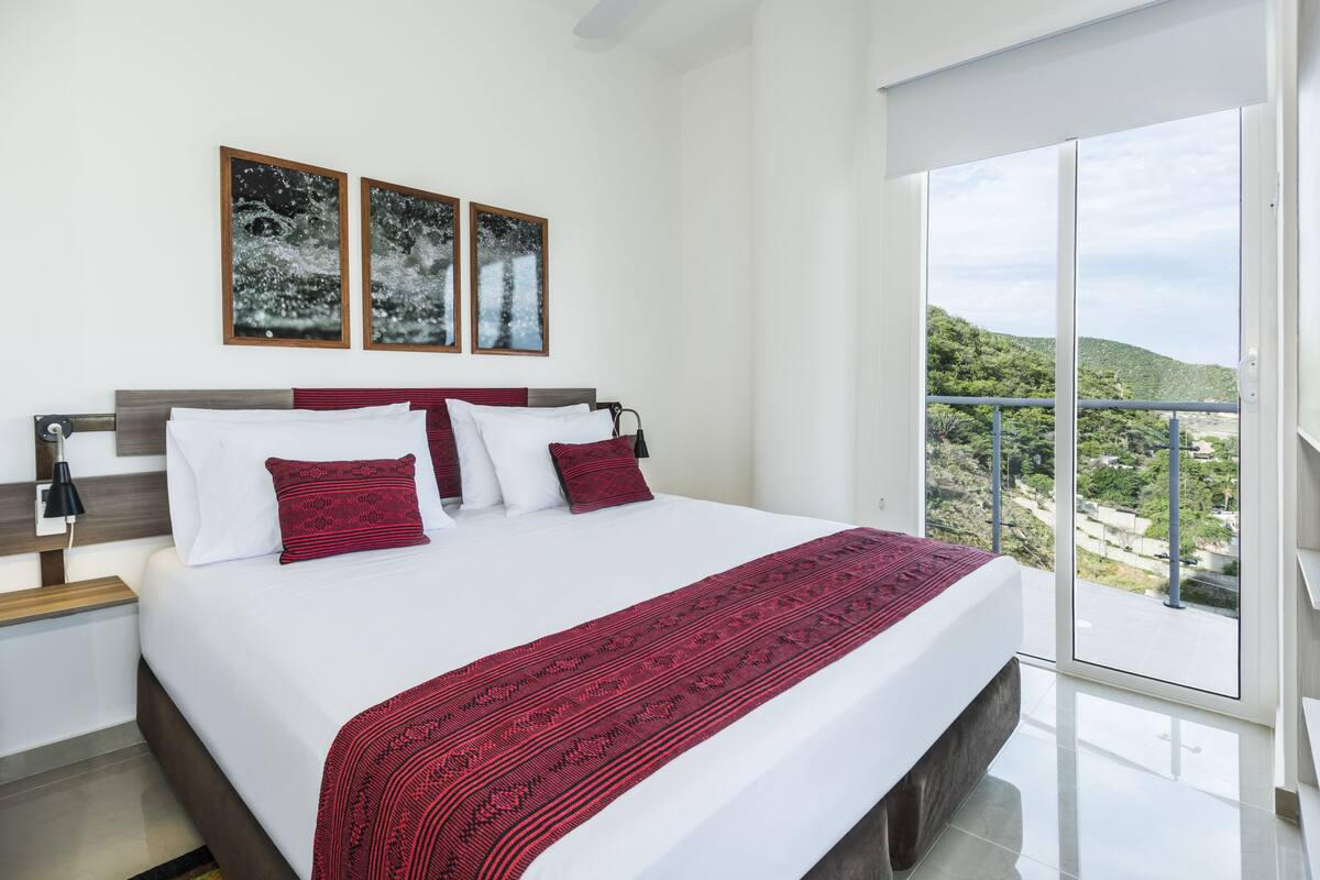 Un dormitorio moderno con una cama grande, ropa de cama blanca, almohadas rojas y una manta decorativa estampada en rojo. Hay dos cuadros en la pared y un balcón con vistas a zonas verdes.