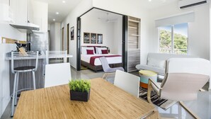 A modern hotel room with a wooden dining table, a bed with red bedding, a white sofa, and a view of greenery outside.