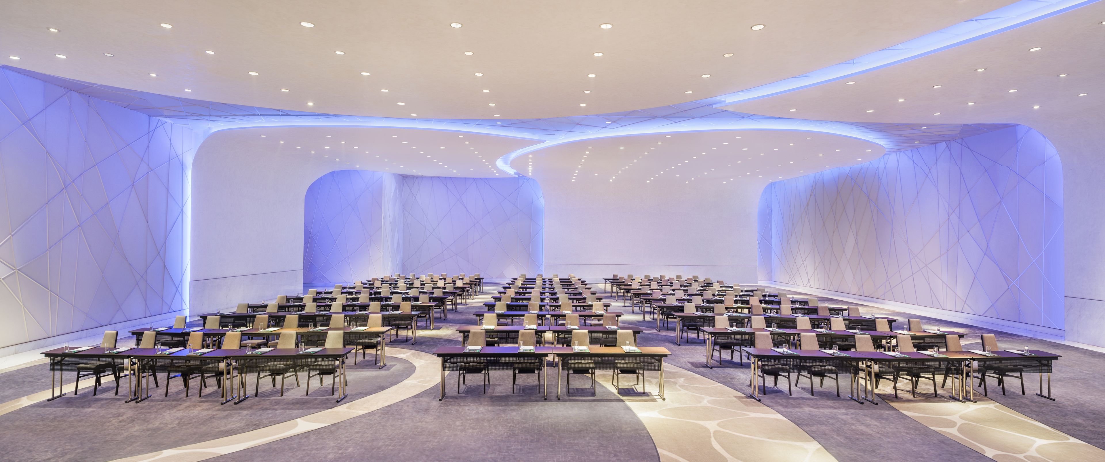 A large conference room with rows of tables and chairs, a high ceiling with recessed lighting, and a modern geometric wall design.