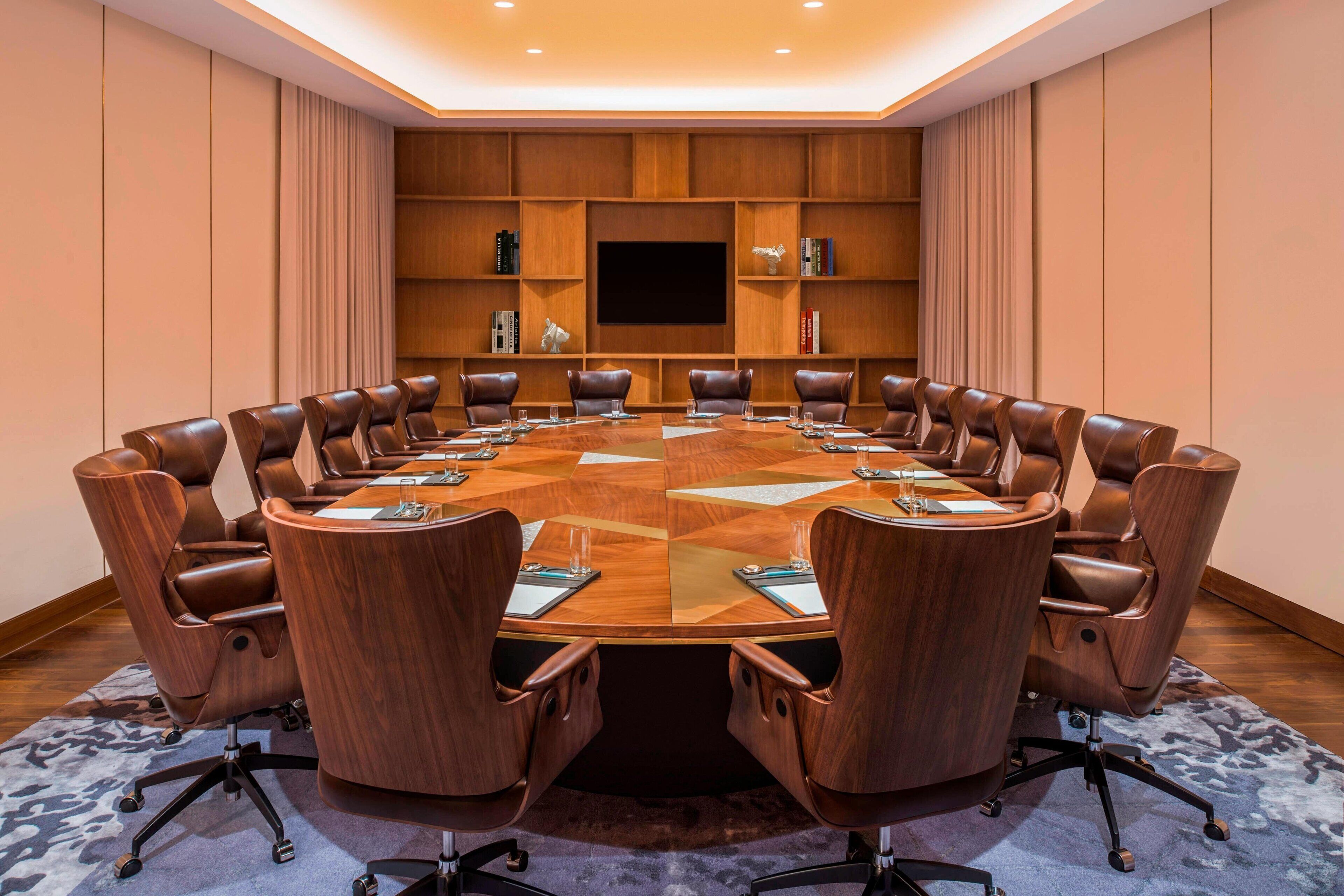 Una sala de conferencias con una gran mesa ovalada, sillas de cuero, un televisor de pantalla plana y estanterías empotradas con libros y objetos decorativos.