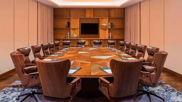Una sala de conferencias con una gran mesa ovalada, sillas de cuero, un televisor de pantalla plana y estanterías empotradas con libros y objetos decorativos.