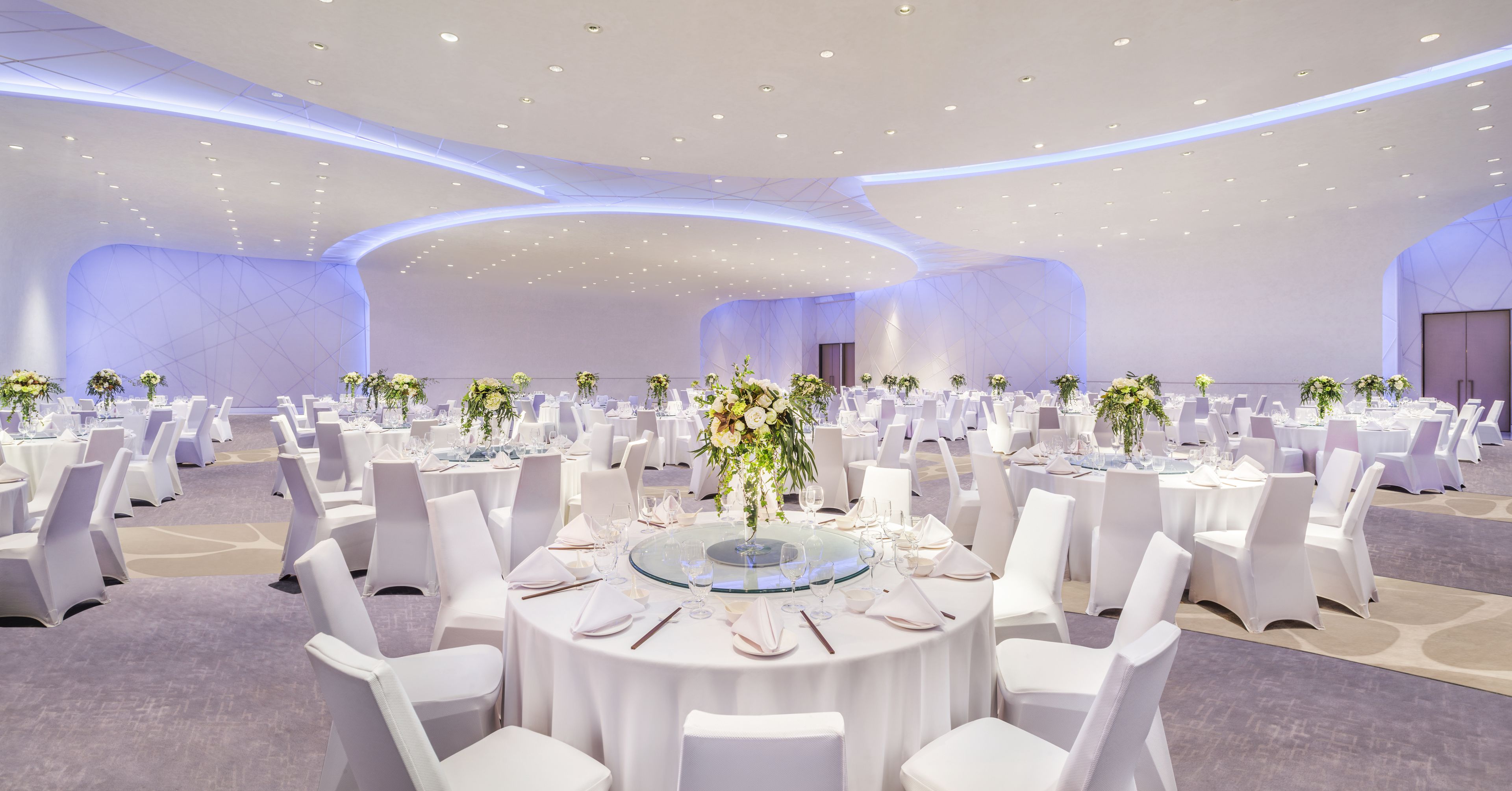 A large, well-lit banquet hall with round tables set for dining, white chairs, and floral centerpieces.