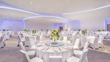 A large, well-lit banquet hall with round tables set for dining, white chairs, and floral centerpieces.