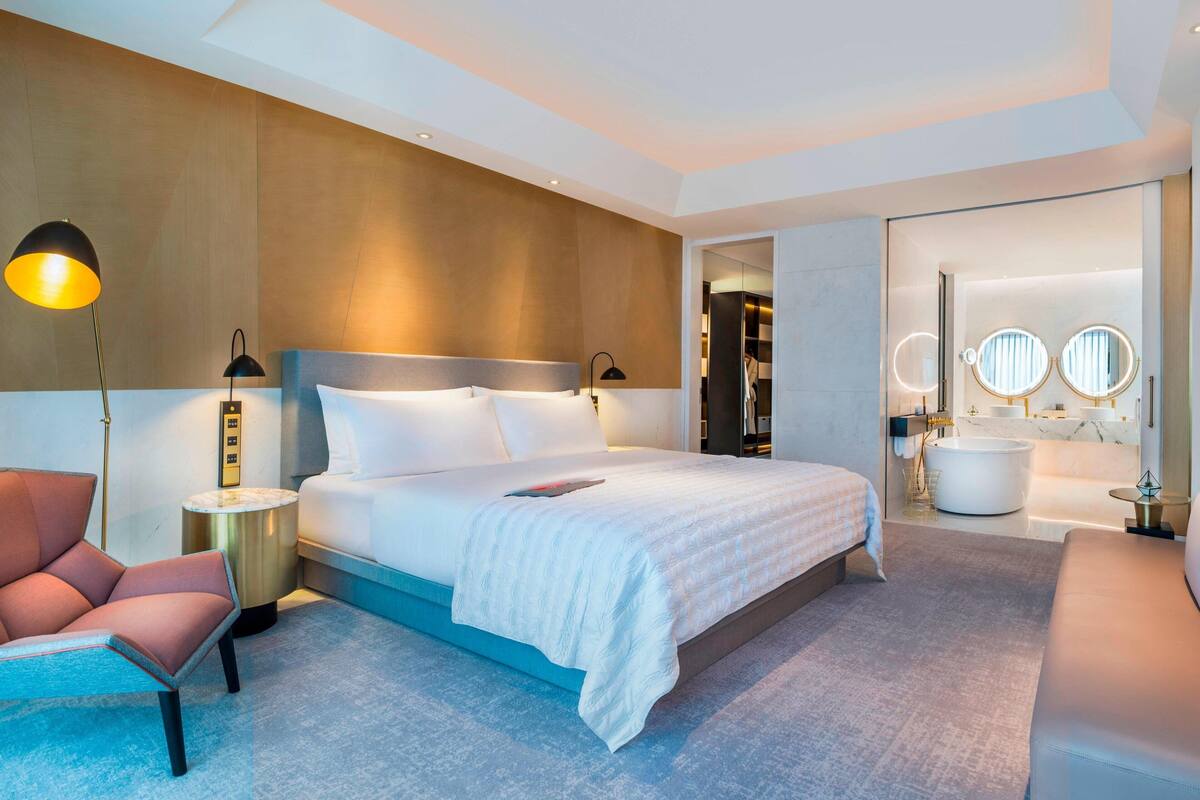 A modern hotel room with a large bed, a sitting area, a vanity with mirrors, and a bathtub.