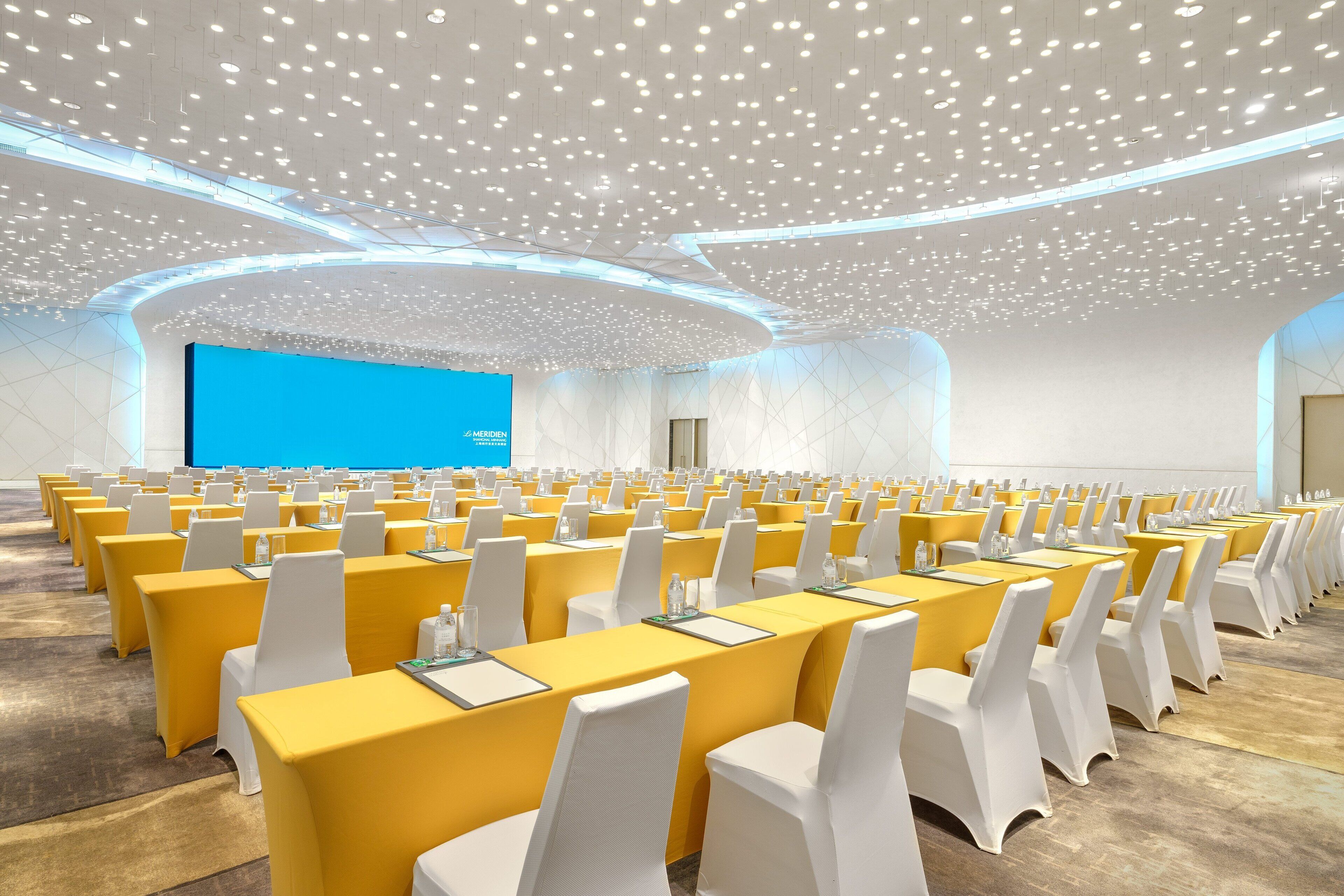 A conference room with rows of yellow tables and white chairs, a large blue screen, and a modern ceiling design.