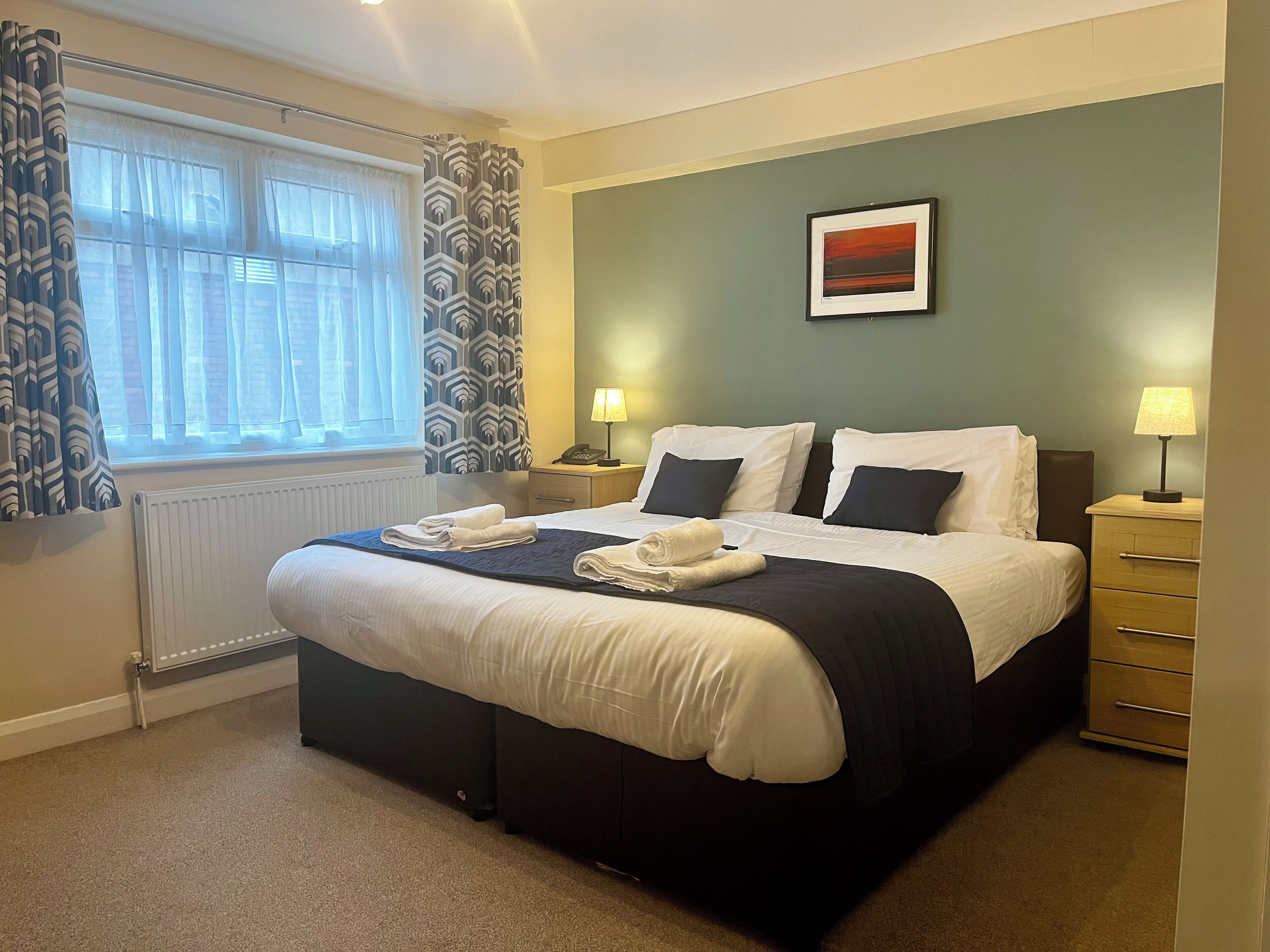 A neatly made bed with white linens and a dark headboard, a bedside table with a lamp, a framed picture on the wall, and a window with patterned curtains.