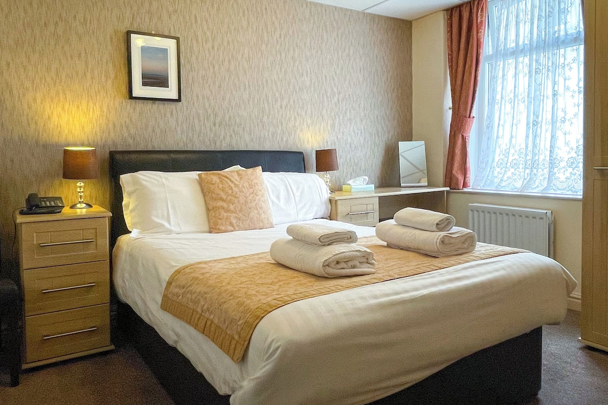 A neatly made bed with a headboard, a bedside table with a lamp, a nightstand, and a window with curtains.