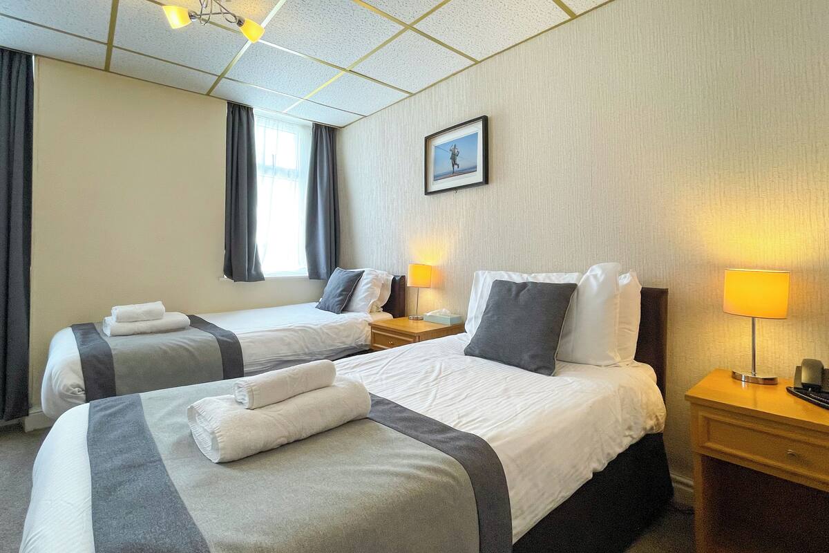 A hotel room with two beds, each with a pillow and a rolled towel. There are bedside tables with lamps and a picture on the wall.