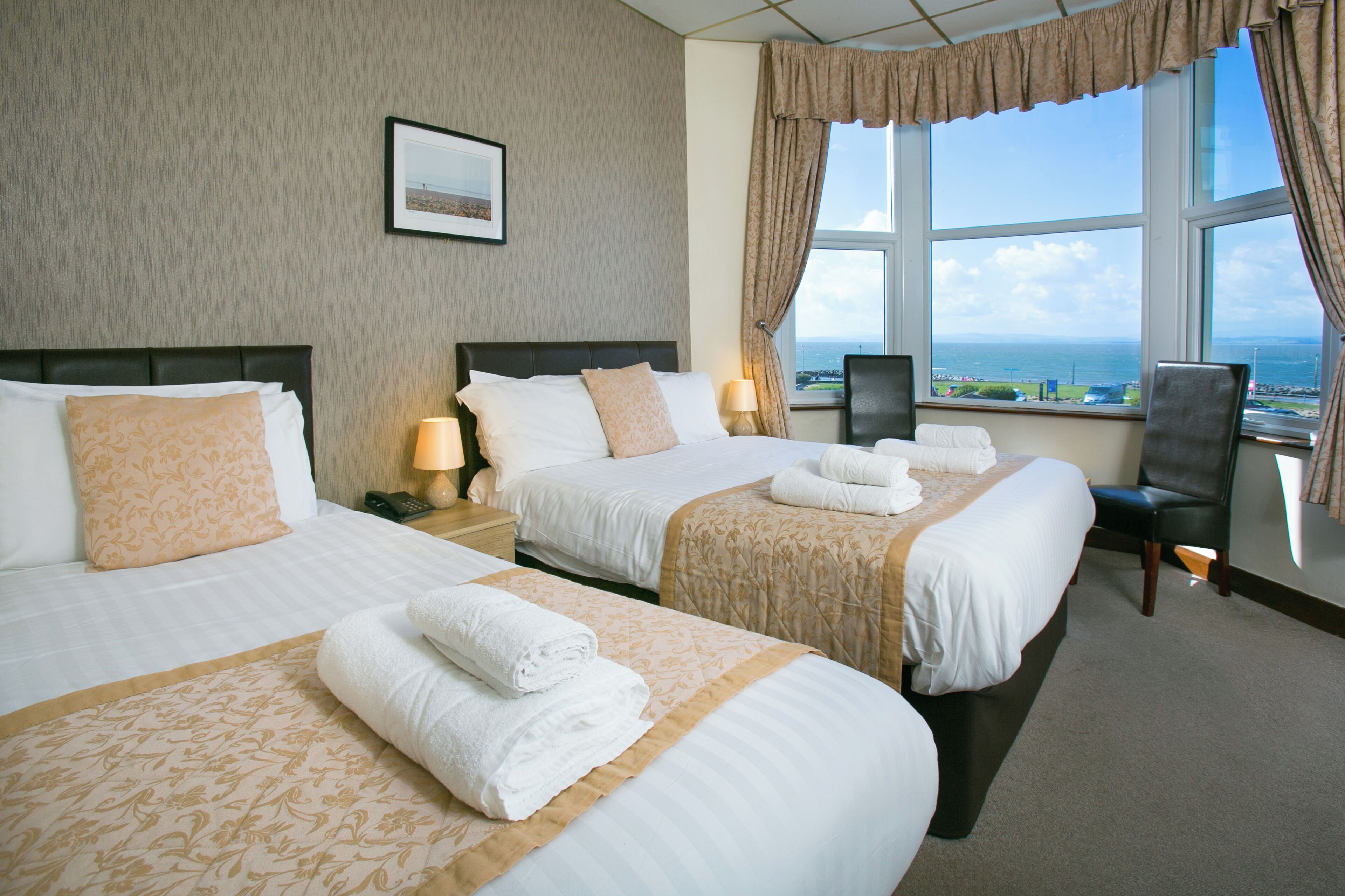 A hotel room with two beds, a desk, a chair, and a large window with a view of the sea.
